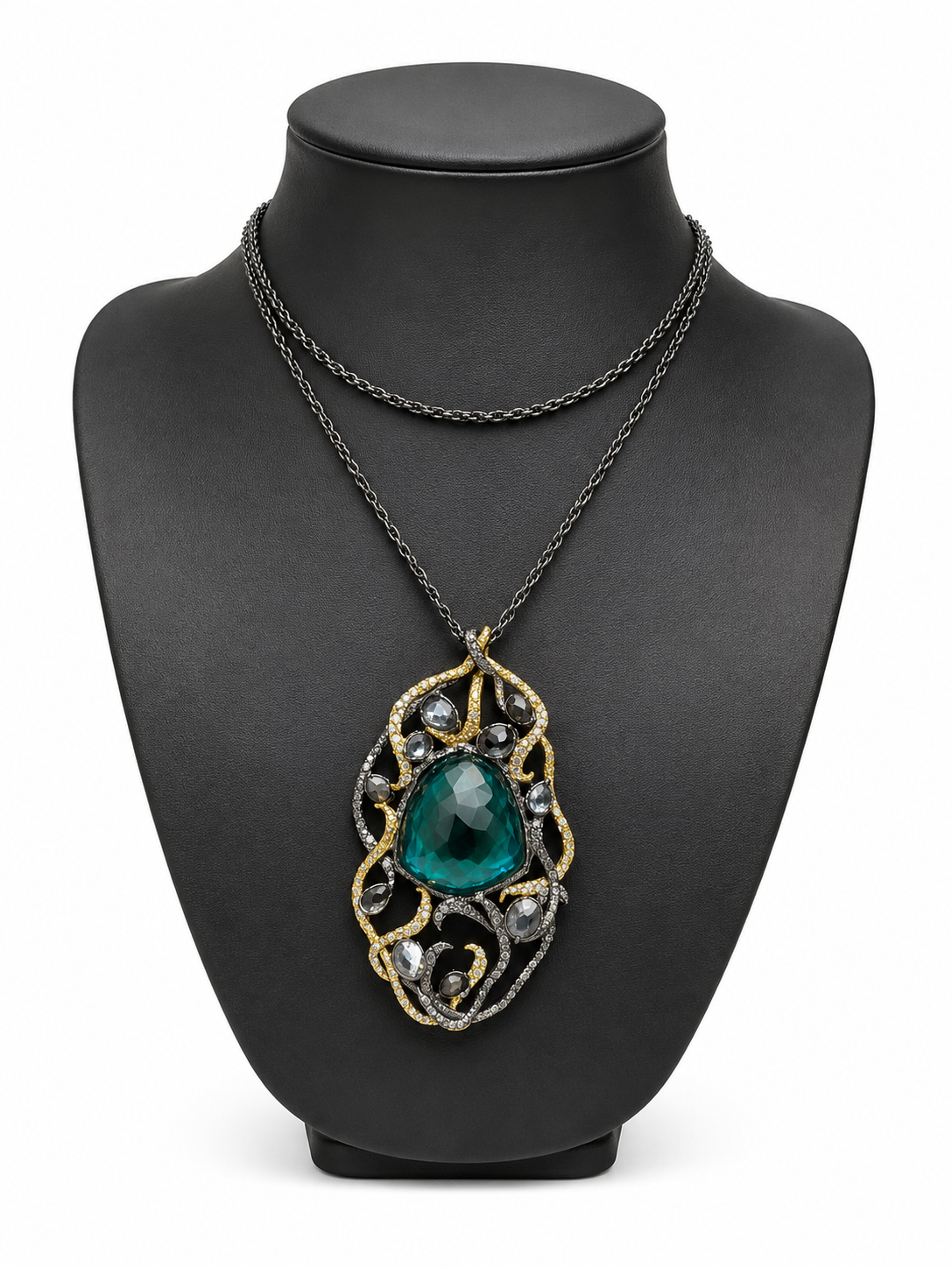 A double-strand necklace displayed on a matte black jewelry bust. The necklace features a dark oxidized chain and a central pendant with a large, faceted teal-green teardrop gemstone surrounded by intricate gold and silver filigree.