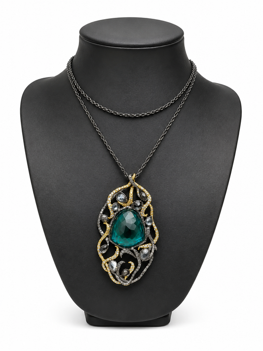 A double-strand necklace displayed on a matte black jewelry bust. The necklace features a dark oxidized chain and a central pendant with a large, faceted teal-green teardrop gemstone surrounded by intricate gold and silver filigree.