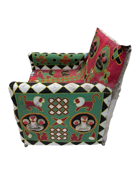 A small upholstered armchair with a highly decorative, multicolored patchwork design featuring green, red, black, white, and gold patterns, including stylized dogs, paw prints, and geometric motifs, shown against a plain white background.