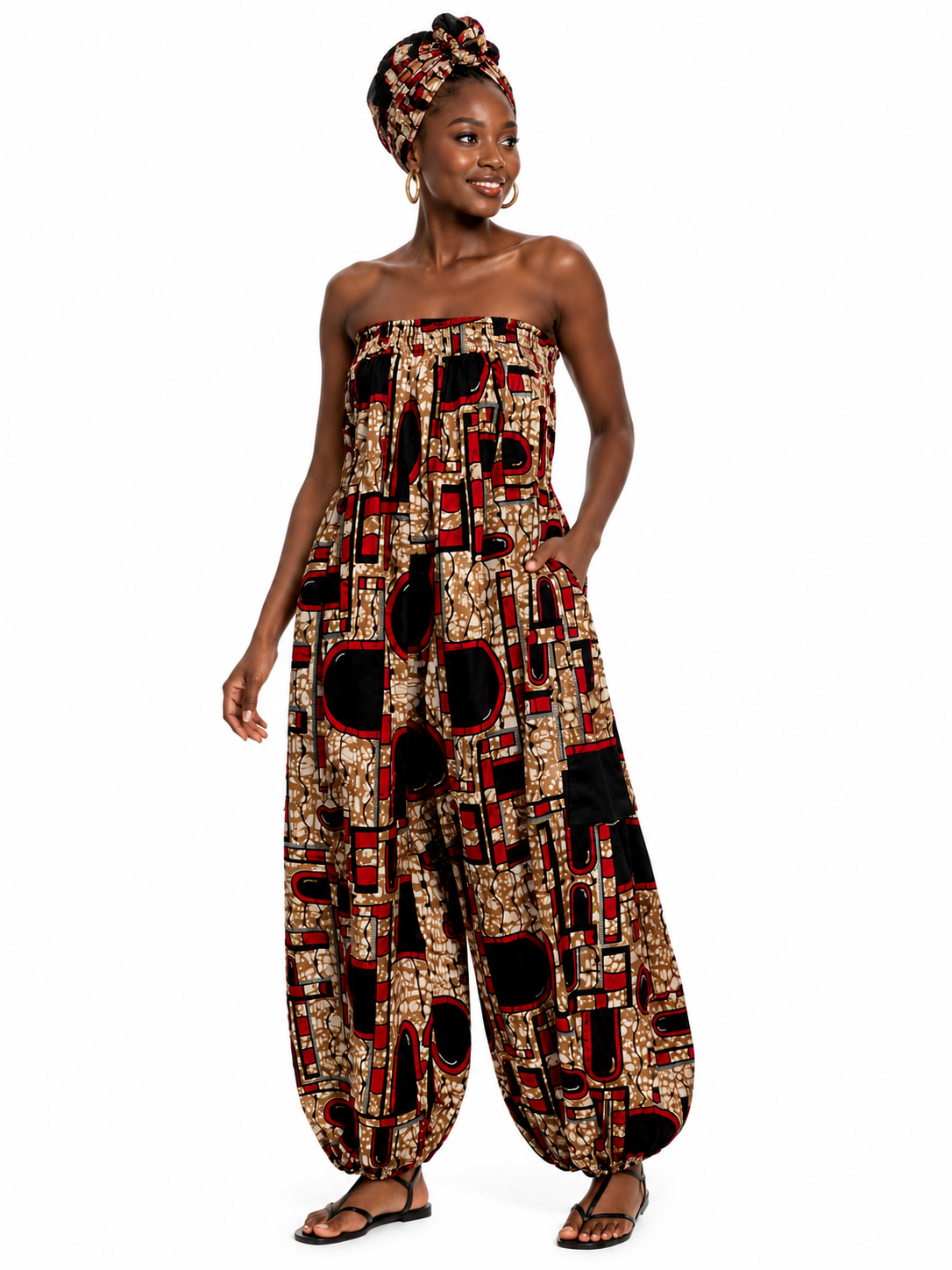 A woman modeling a strapless harem-style jumpsuit with a bold geometric pattern in black, red, and beige, paired with a matching headwrap and flat sandals, set against a plain white background.