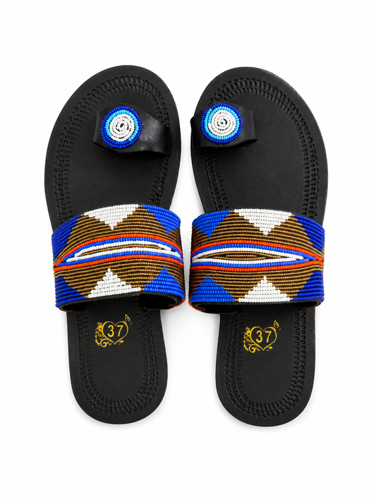 A pair of black slide sandals with wide beaded straps featuring geometric patterns in blue, brown, white, and orange. Each sandal has a small circular beaded toe loop and a gold monogram logo printed on the footbed.